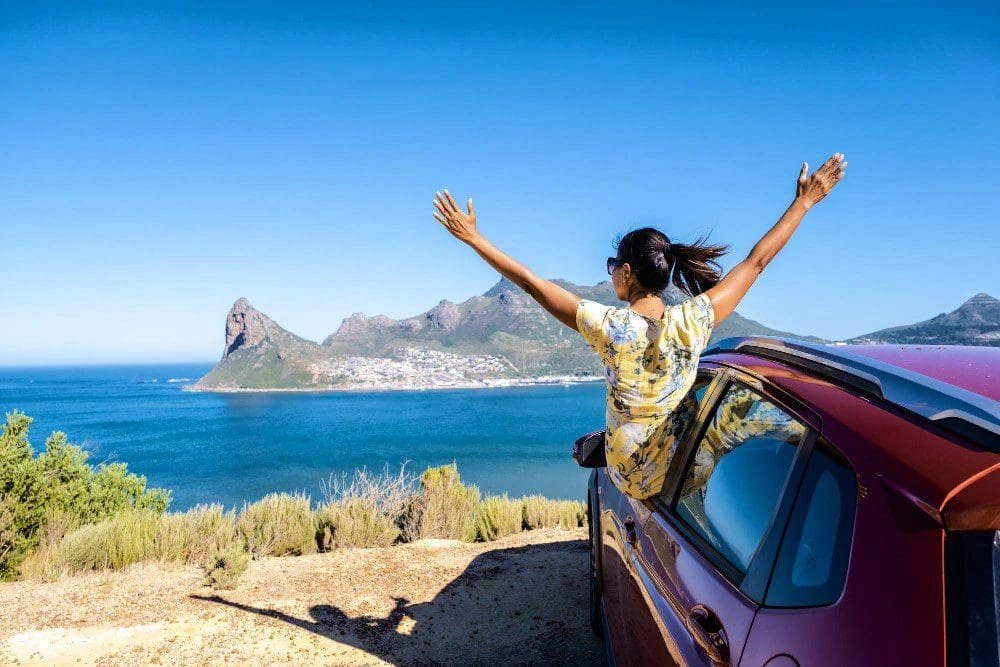 A-woman-enjoying-a-road-trip-on-Chapmans-Peak-Drive-in-Cape-Town-with-her-hands-up-gazes-out-over-the-ocean-from-the-car-window in africa